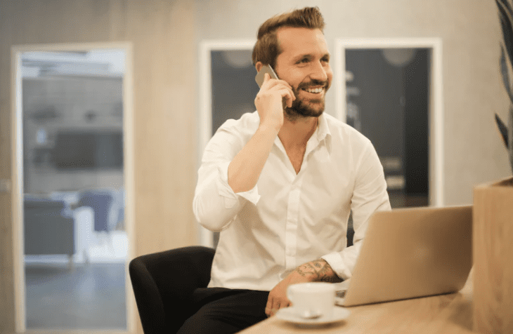 white male with a beard holding a phone to his ear sits at a desk with a laptop and cup of tea